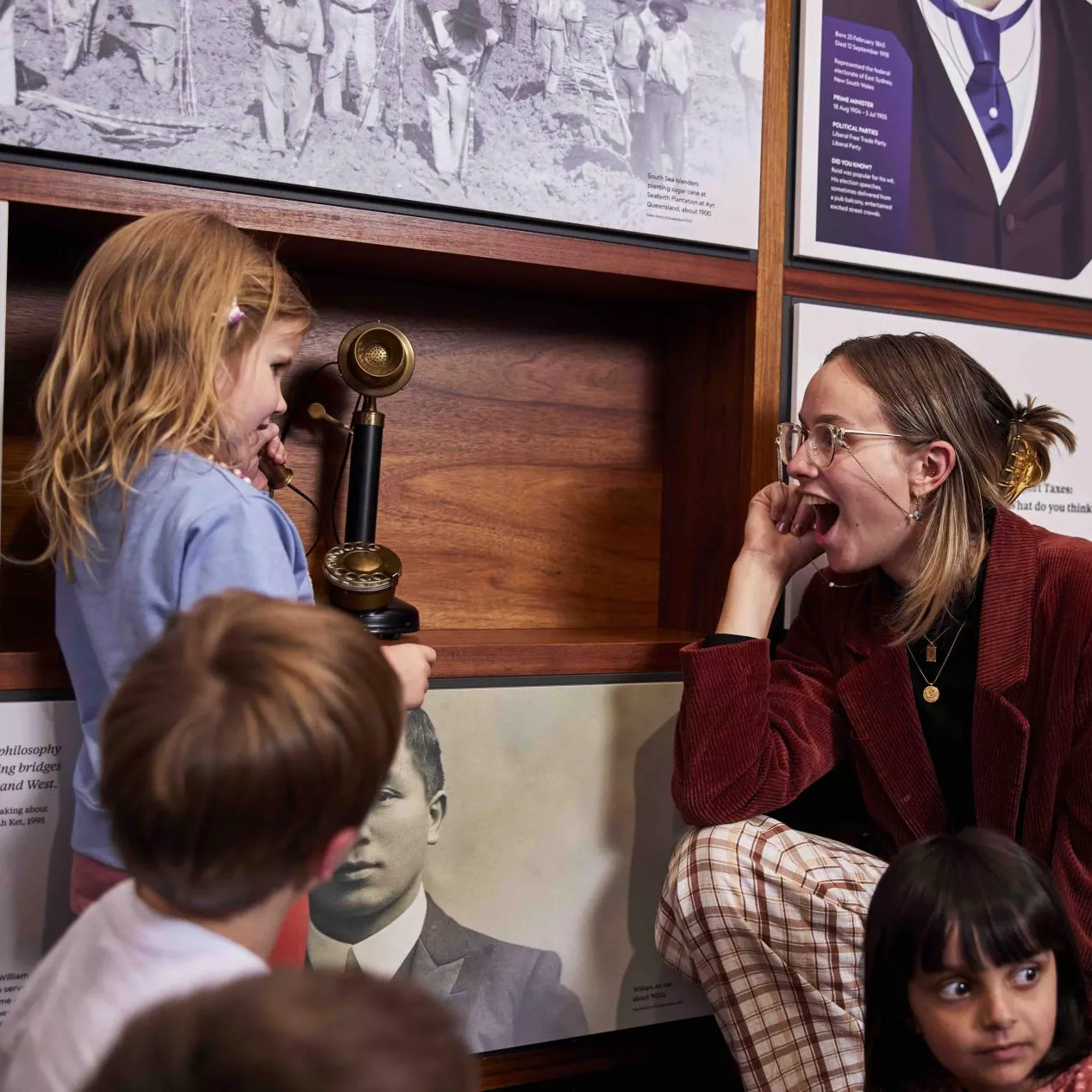 A woman smiles at a child holding a telephone to her ear in a museum exhibition with panels about prime ministers. 