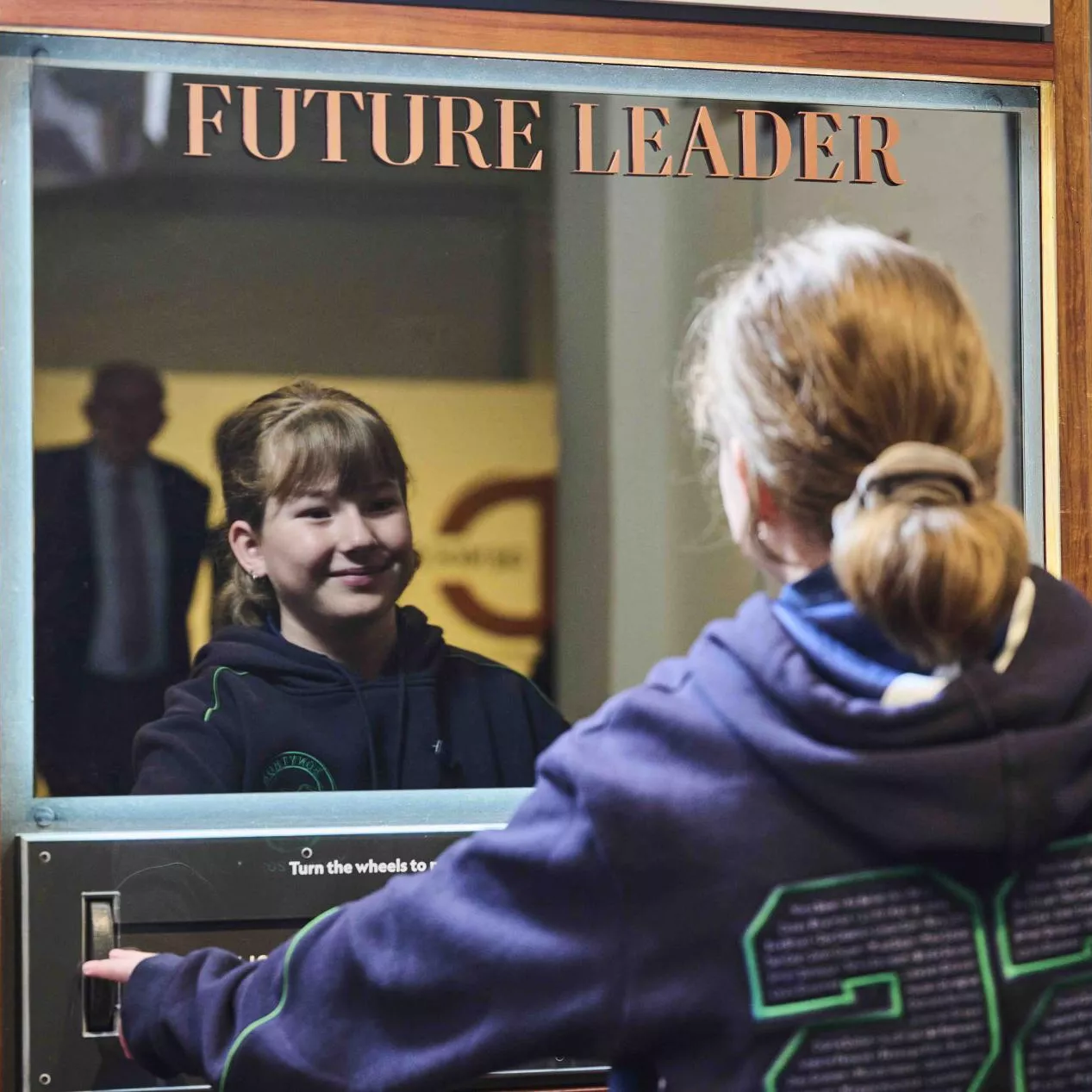 A primary school girl wearing her school jumper looks into a mirror with the words future leader on it. 