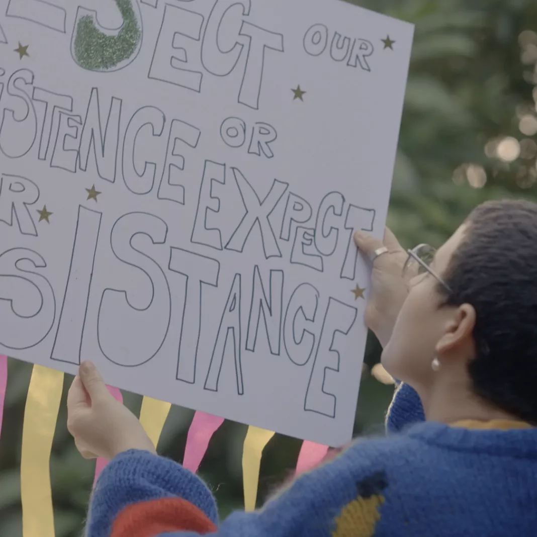 A person holds up a protest placard with the label "Respect our existence or expect our resistance"