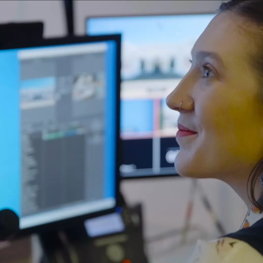 A woman looks off to the side and some screens and other technology are out of focus in the background