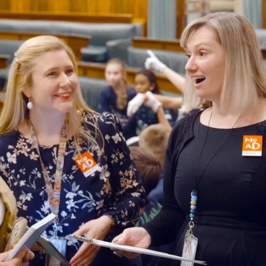 Two people stand holding clipboards, smiling and talking to a person about old parliament house's chamber of representatives.