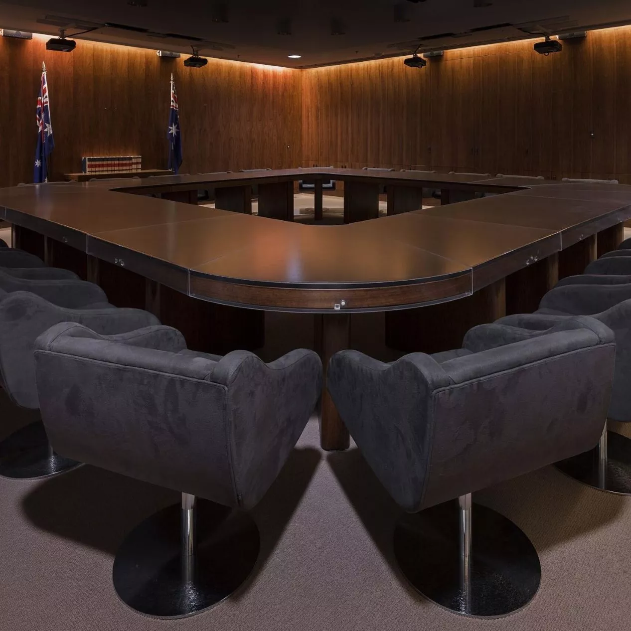 A dark room with heavy curtins and a large, heavy oval shaped table with a hole in the middle surrounded by large grey chairs. 