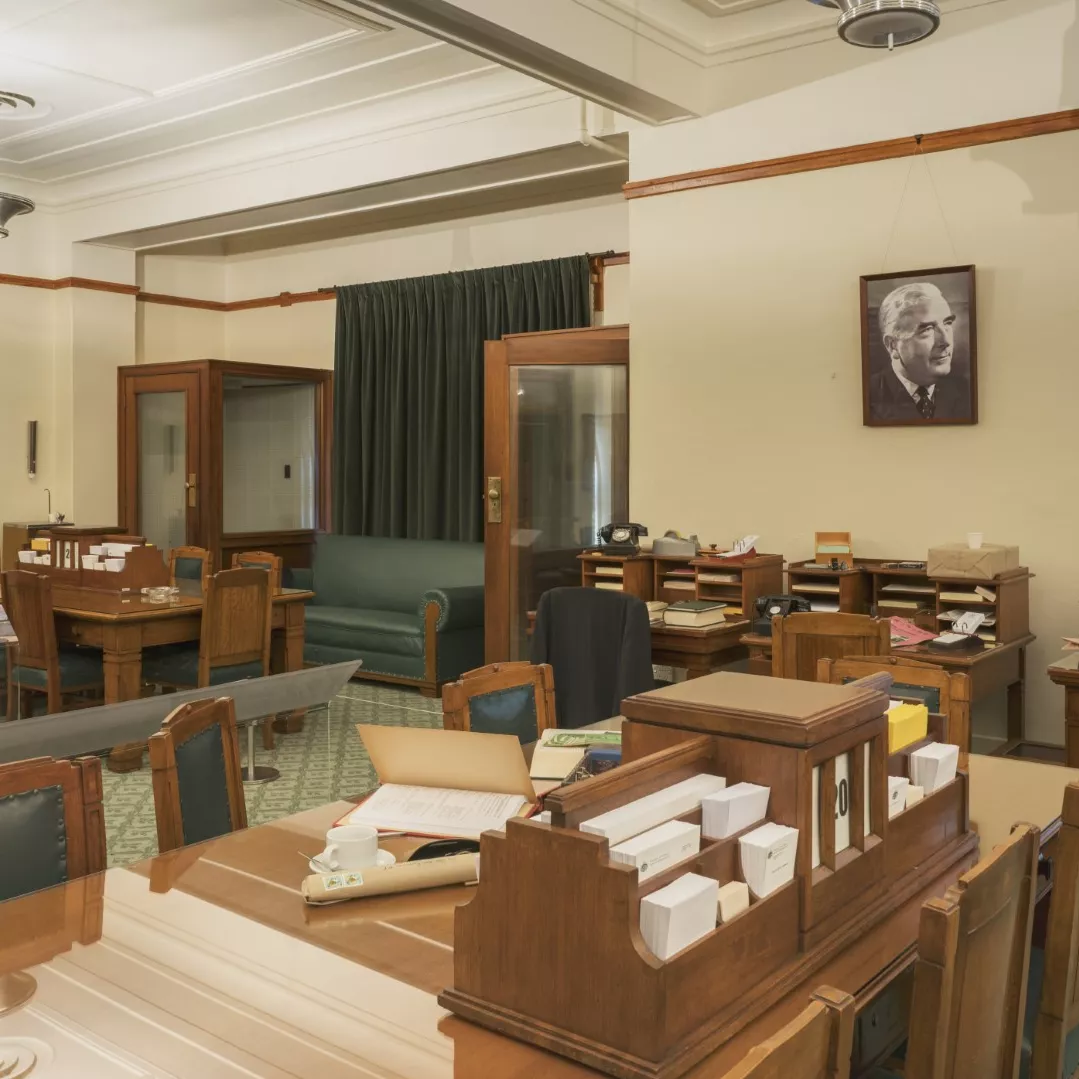 This colour photograph shows the Government Party Room re-created to evoke the atmosphere of 1964. There are two large meeting tables surrounded by chairs, three small desks, and two telephone booths. A large portrait of Prime Minister Robert Menzies is on one wall. The surfaces of the meeting tables and desks are covered with stationery, papers, telephones and ashtrays. 