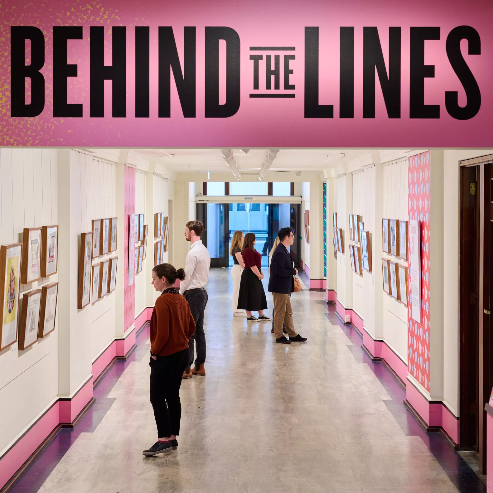 People look at artwork hanging on walls in a gallery, with the words Behind the Lines presented above the entrance in black letters on a pink background.