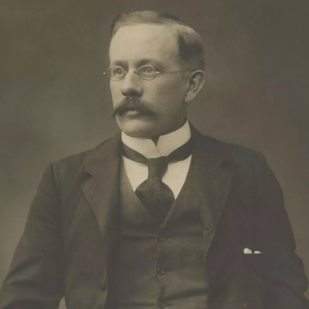 A photographic portrait of Hugh Mahon, wearing glasses and a three-piece suit.