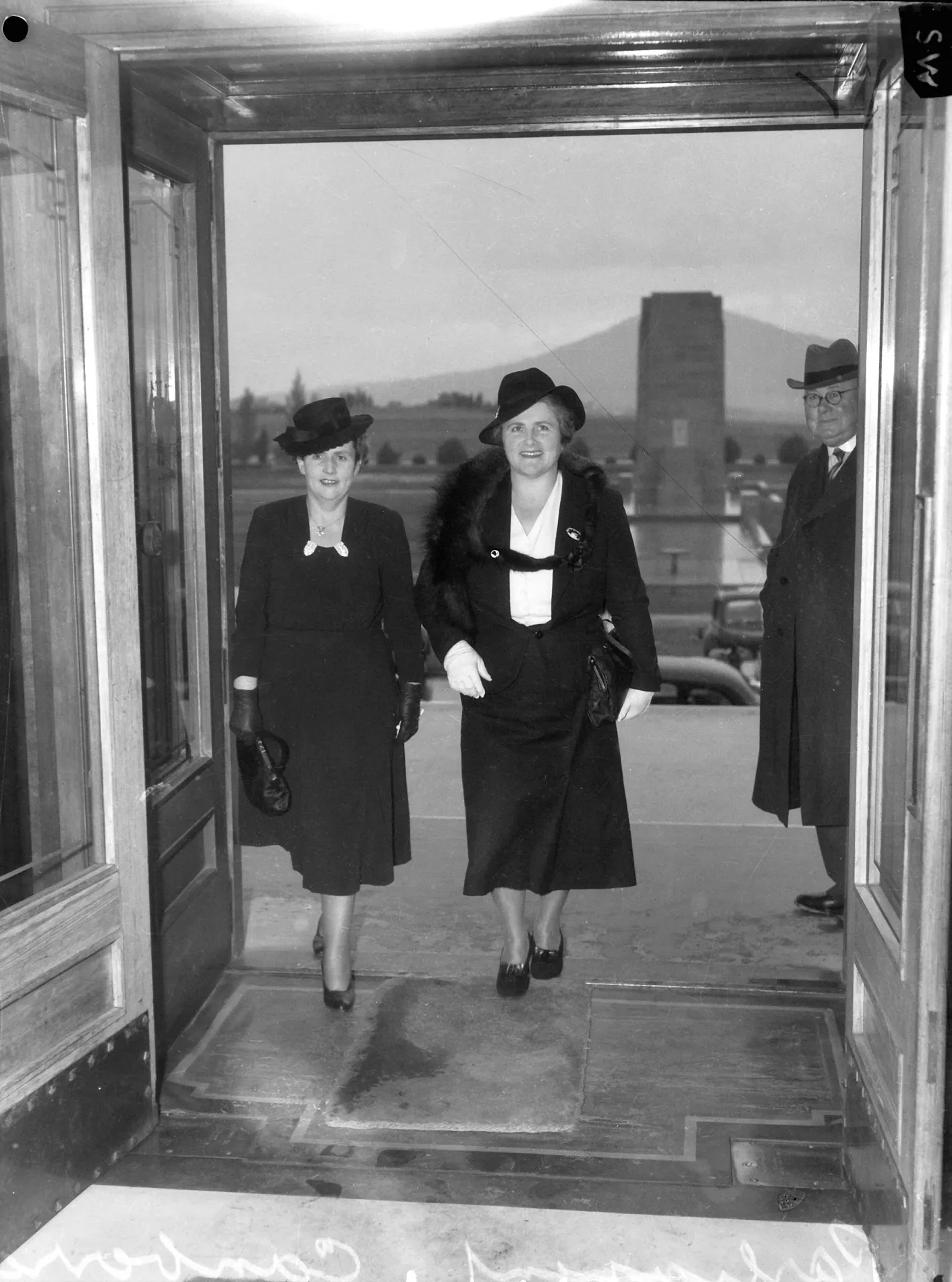 Dorothy Tangney and Enid Lyons walk together through the front door of Old Parliament House, smiling.