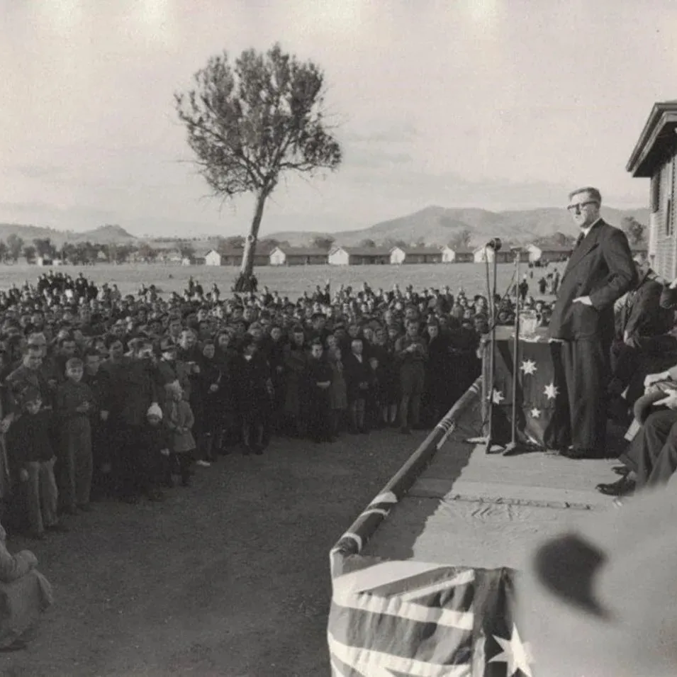 Arthur Calwell stands on a platform with microphones, addressing a crowd of people. In the distance is a row of long buildings.