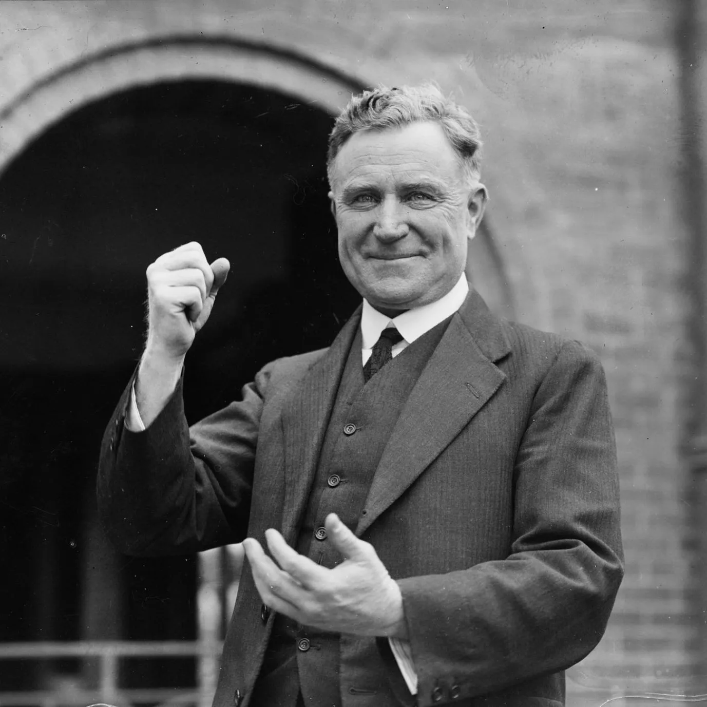 A man, Earle Page, wearing a striped three-piece suit, smiles and poses for a camera with a raised fist.