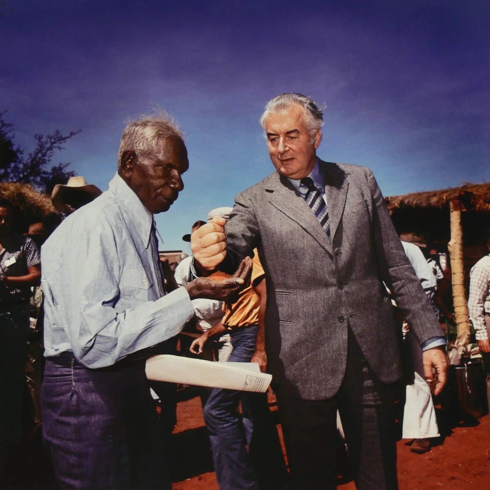 Prime Minister Gough Whitlam pouring earth through Vincent Lingiari’s hand while people look on.