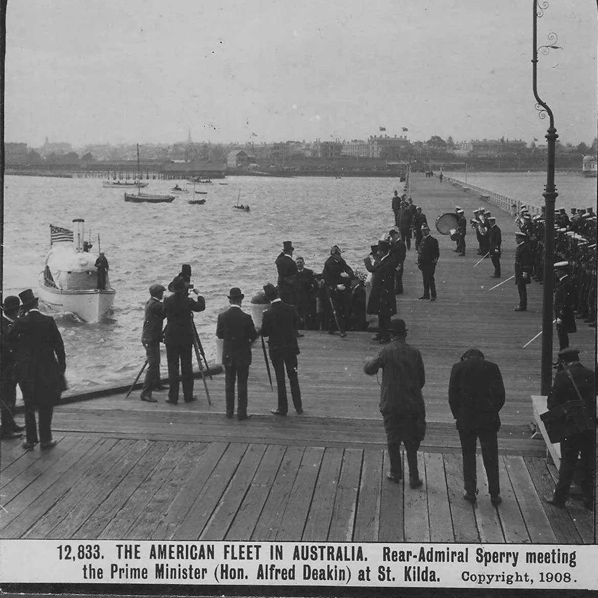 An elevated view from above and behind small clusters of people who stand on a wooden pier. They are looking out at a boat that is headed towards the pier.  The label reads "The American Fleet in Australia. Rear-Admiral Sperry meeting the Prime Minister (Hon. Alfred Deakin) at St. Kilda." 1908.