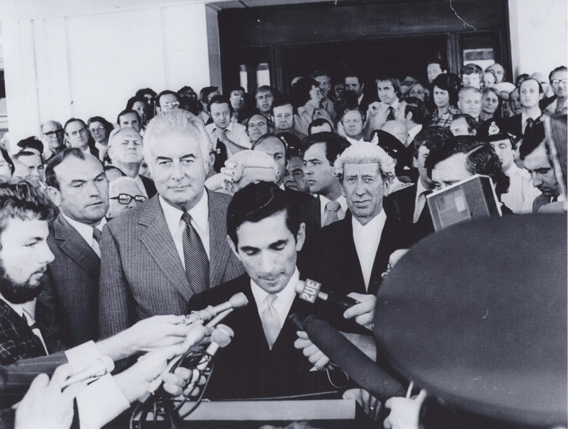 A black and white photo of a Gough Whitlam on the steps of Parliament House surrounded by a crowd of men in suits. One man at the front speaks from a lectern as journalists put their microphones in his face. A man in the background wears a barrister's wig and looks at the camera. 