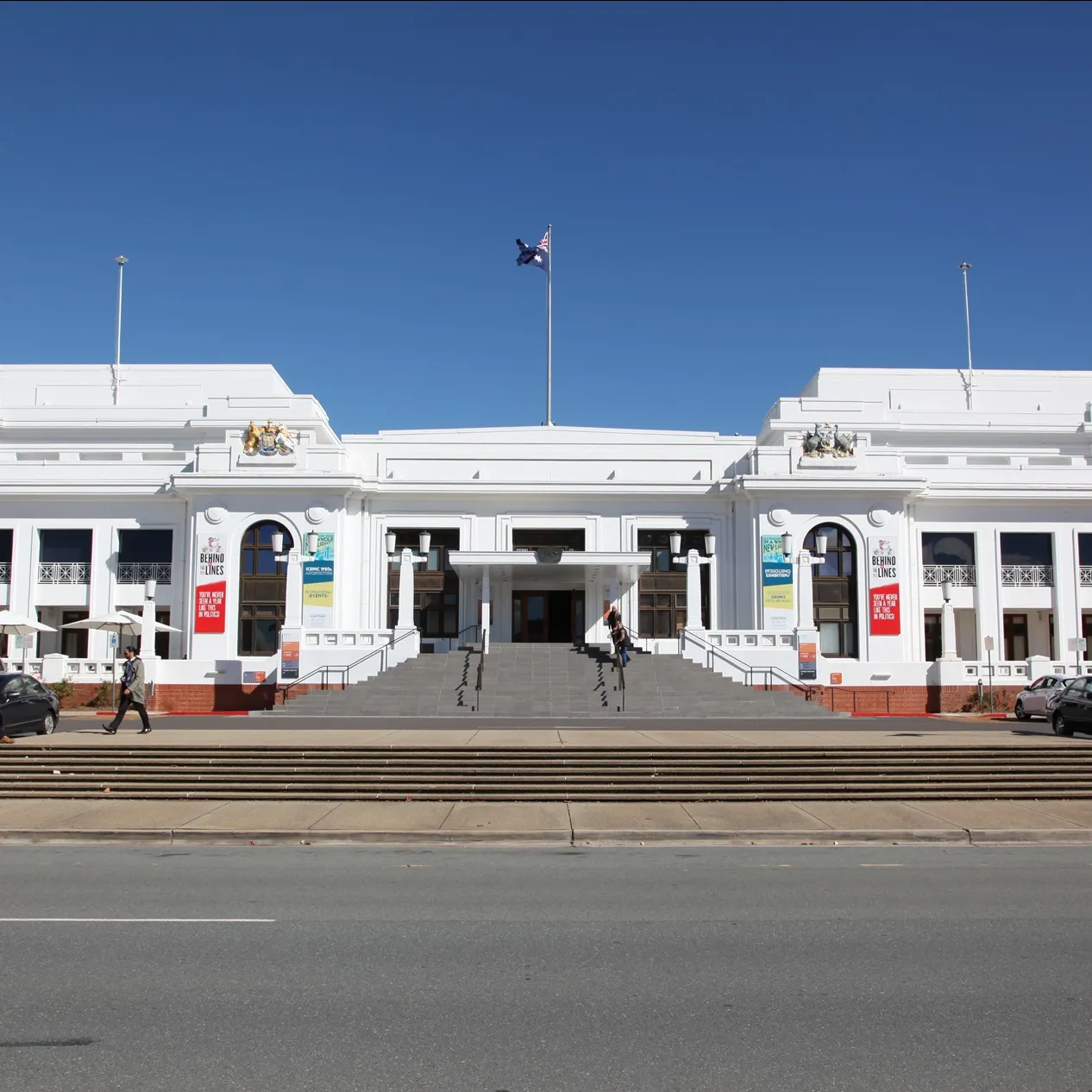 Front of Old Parliament House, flying the Australian flag and two Aboriginal flags, with people standing at the front stairs.