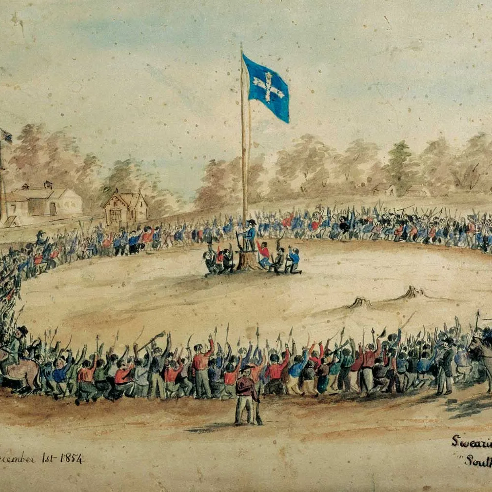 A painting of a large group of miners gathered in a circle around a flagpole, kneeling and raising hands and objects into the air, as the Eureka flag is raised. The title reads 'Swearing allegiance to the "Southern Cross"' and dated 'December 1st 1854'.