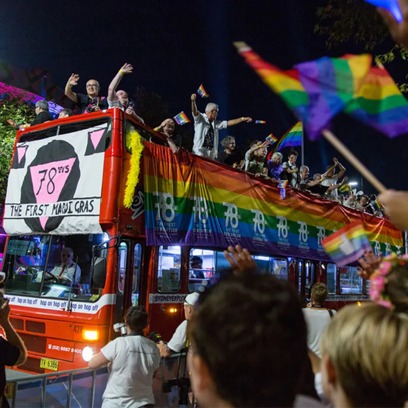 A red double-decker bus with rainbow banners displaying the words '78ers: the first mardi gras' with people on top of the bus waving to crowds on each side of the street.