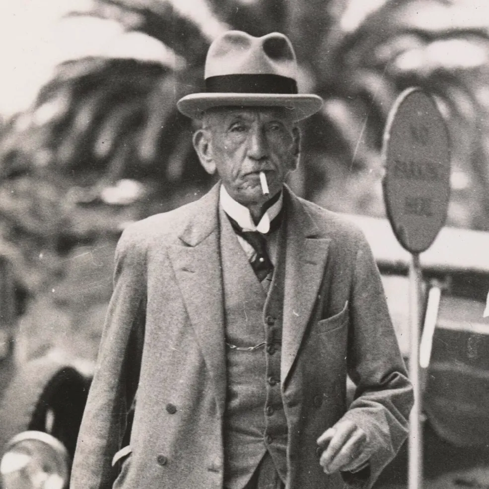 Detail of William Hughes walks down the street wearing a hat and three-piece suit, smoking a cigarette.