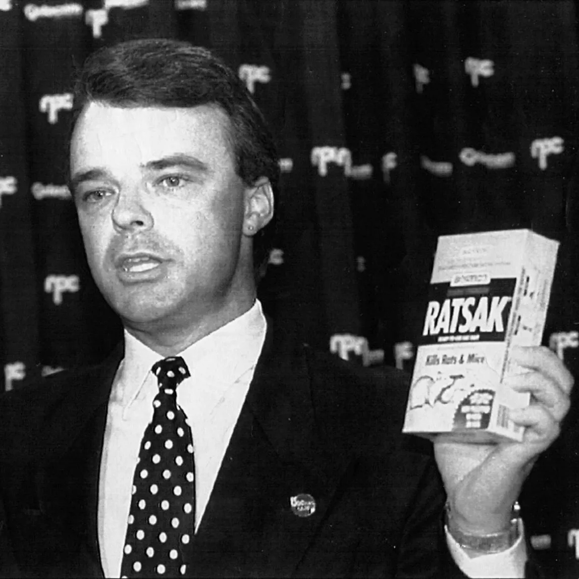 Dr Brendan Nelson, wearing a suit and spotted tie, holding a 'Winfield' cigarette box in one hand and a 'Ratsak' box in the other.