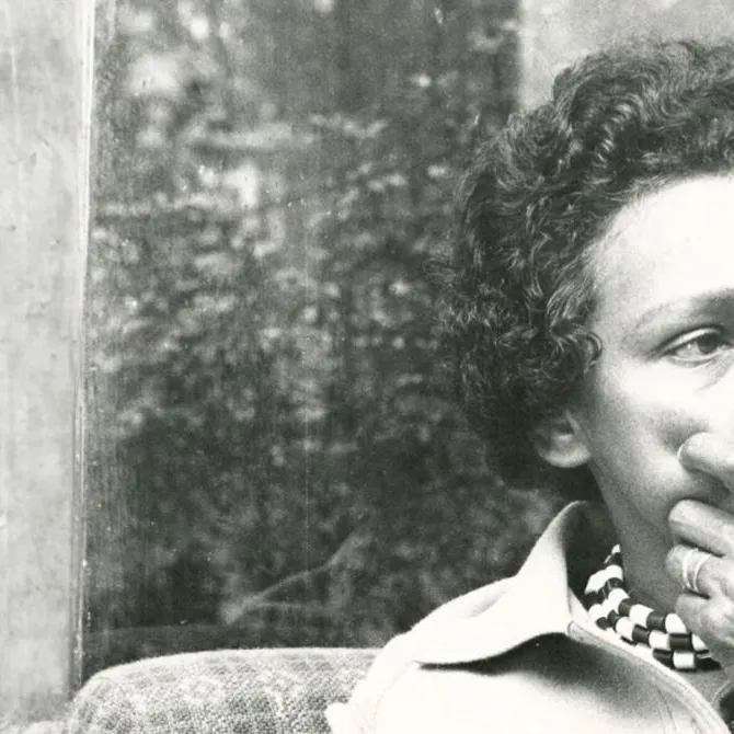 Faith Bandler, sitting on a plaid upholstered chair, holding a hand up to her face.