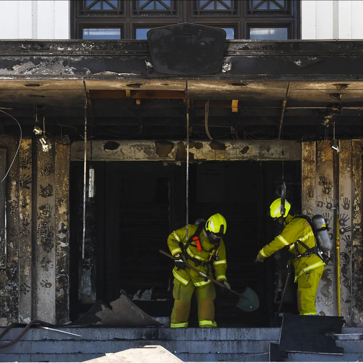 Two firefighters dressed in yellow hi-vis clothing and protective equipment, hold shovels and assess the fire damage to the front entrance of Old Parliament House.