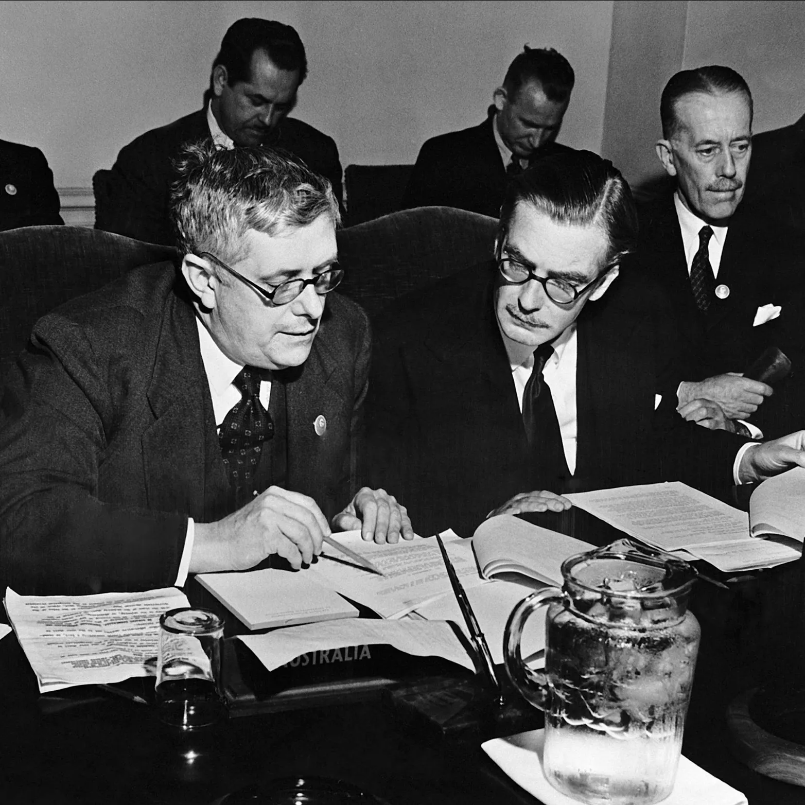 Dr Herbert Evatt sits next to Anthony Eden, reading through typed paper documents, at a table with a glass jug of iced water, and a nameplate labelled 'Australia'.