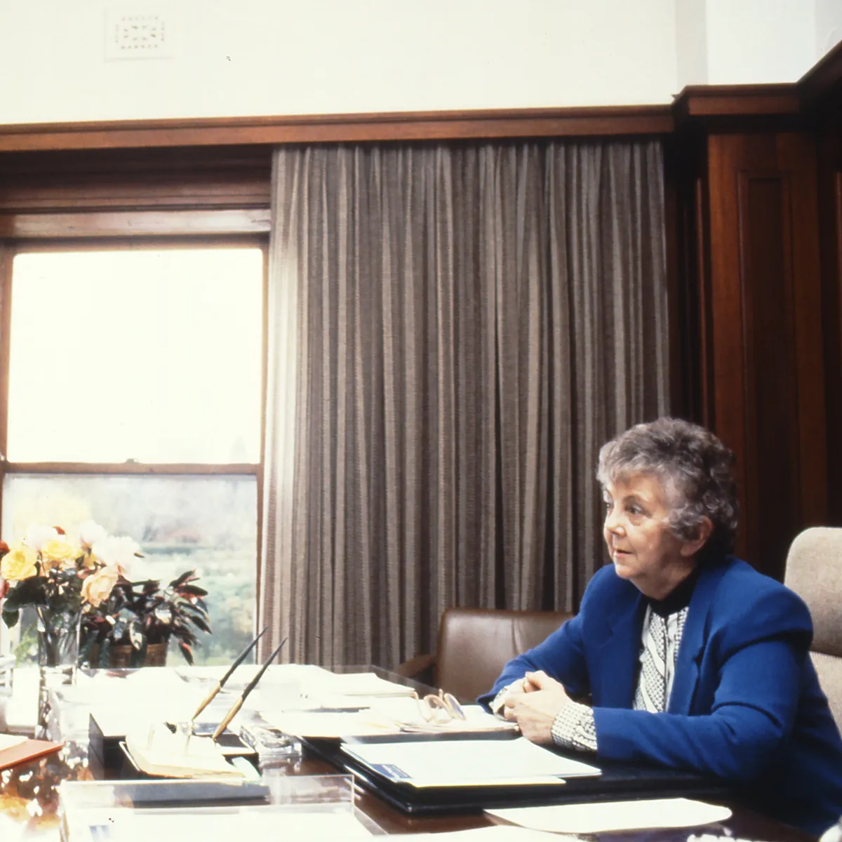 This professional colour photograph shows Principal Private Secretary John Porter and Speaker Joan Child sitting across from each other at the Speaker’s desk. John is wearing a dark suit, white shirt and dark tie and Joan is in a white blouse and vibrant blue jacket. They are discussing the daily program for the House of Representatives Chamber. There is a vase of roses, stationery and paperwork on the desk. In the background the curtains have been drawn to reveal a large east-facing window.