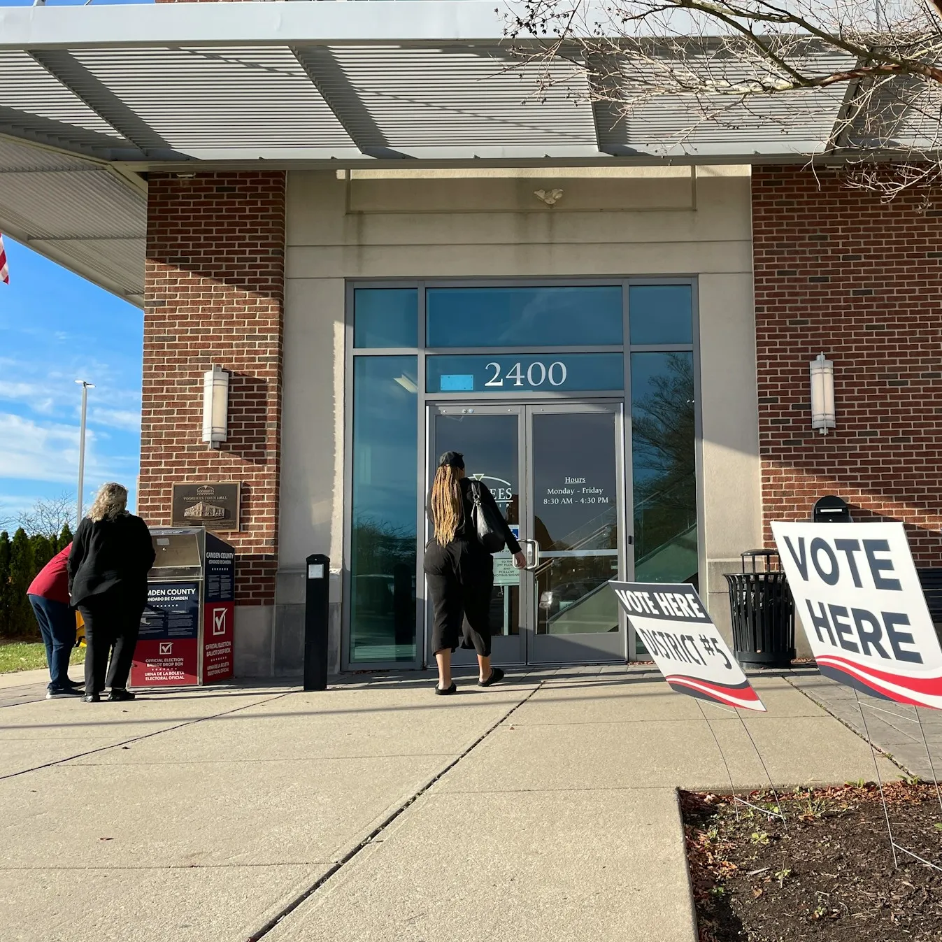 The front of a building with signage that reads 'Vote here', 'District #5' and 'District #12'. One person enters the building, whilst two others gather around a box that reads 'Official election ballot drop box'.