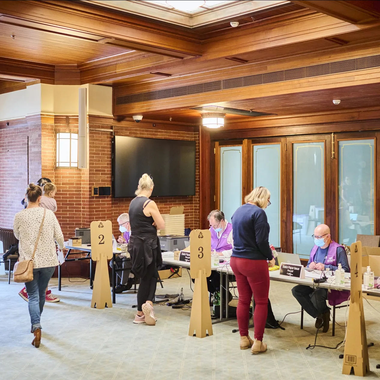 Four Australian Electorial Commission staff sit in a row at a long table wearing face masks and purple vests, and people are lined up, one in front of each staff member, to collect their ballots.