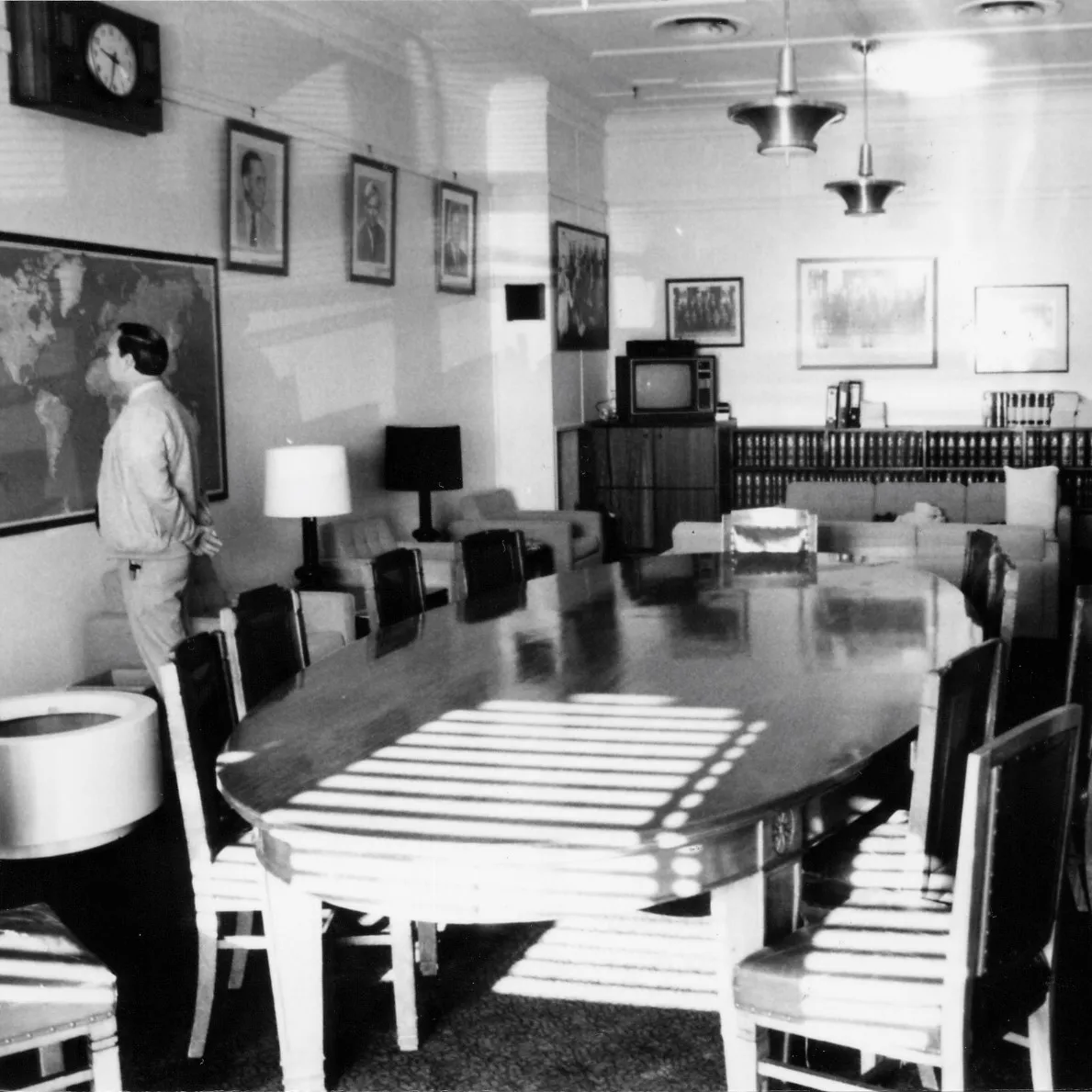 A large oval meeting table and chairs dominate the centre of the room. Easy chairs with side tables and lamps are arrayed along the sides and two sofas are arranged in a seating area towards the back of the room. A sideboard with books, a television and sound system stretches along the back wall. There is a framed map on one wall and a notice board on the opposite. Portraits of leaders and formal and informal photographs of the party crowd the walls