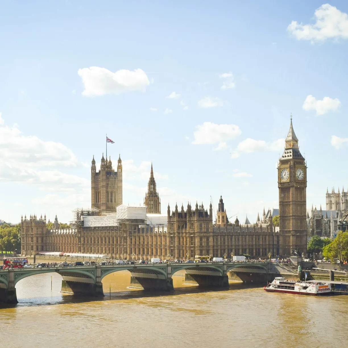 Big Ben and the Palace of Westminster, as viewed from across the river, with red double-decker buses and pedestrians crossing the bridge nearby.