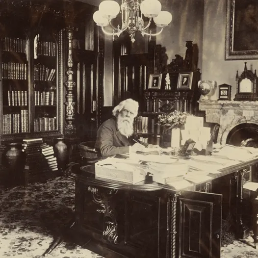 Sir Henry Parkes sits at his desk, with papers and books scattered throughout the room. 