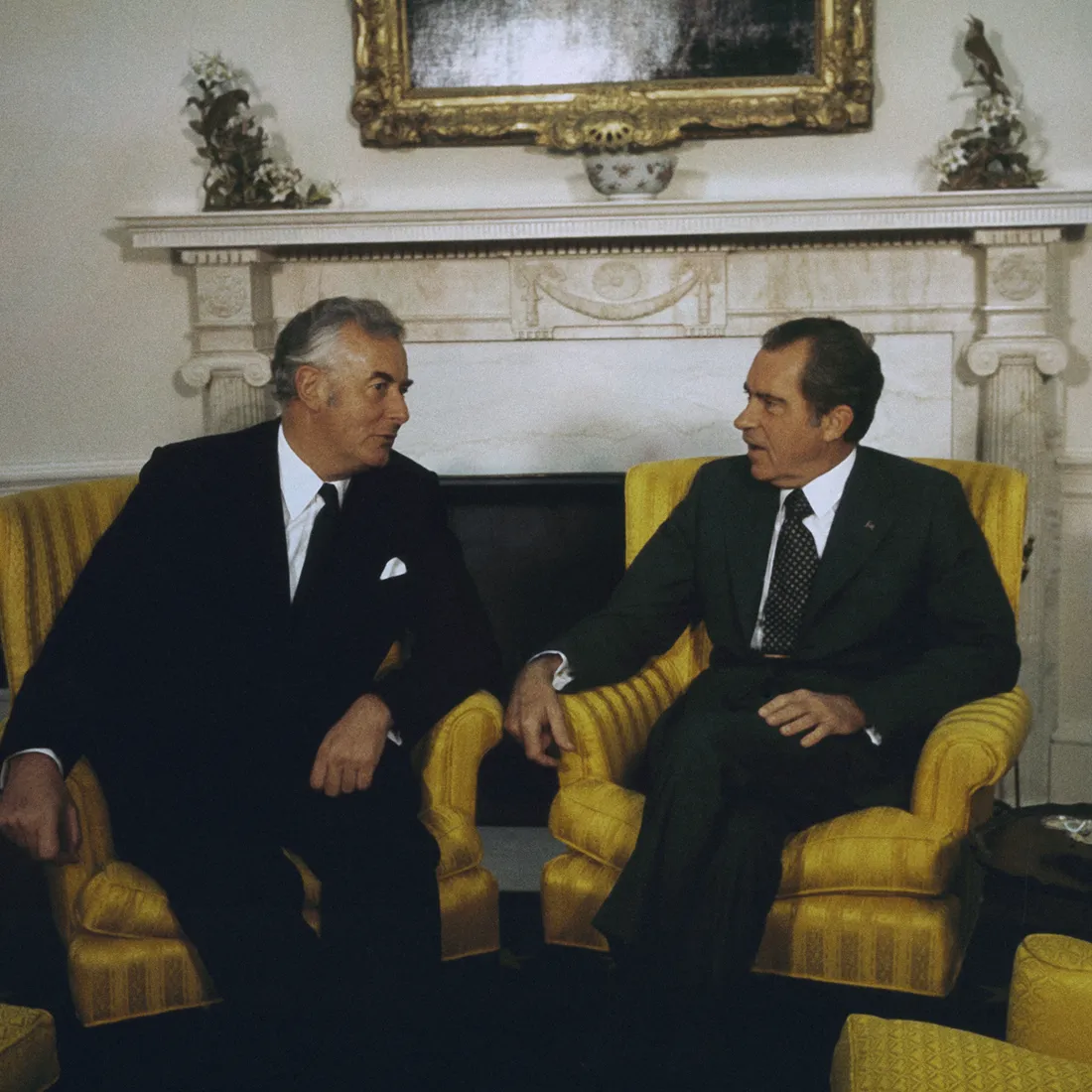 Richard Nixon and Gough Whitlam sit in yellow chairs in the Oval Office facing each other and talking.