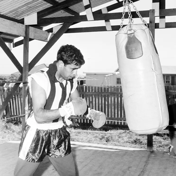 Lionel Rose is dressed in boxing gear. He is looking at a punching bag. 