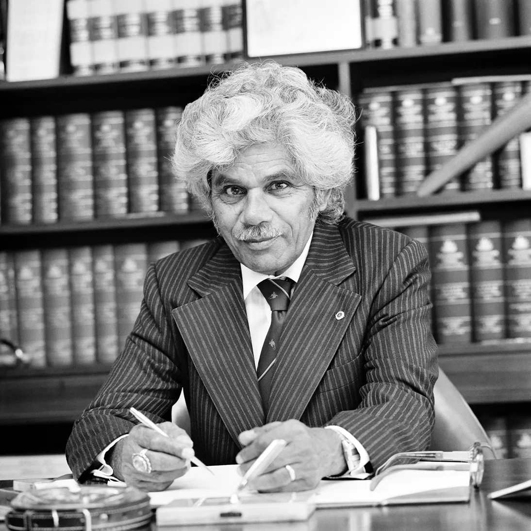 Neville Bonner is sitting at his desk wearing a suit, holding a pen. There are books on bookshelves behind him.