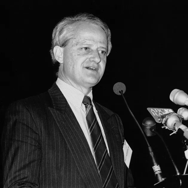 Phillip Ruddock is speaking into an array of microphones. 