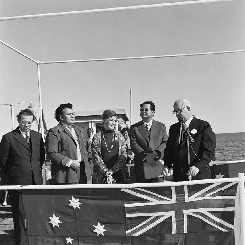 Reg Withers stands behind an Australian flag at a microphone. There are four people to his left. Water is in the background.