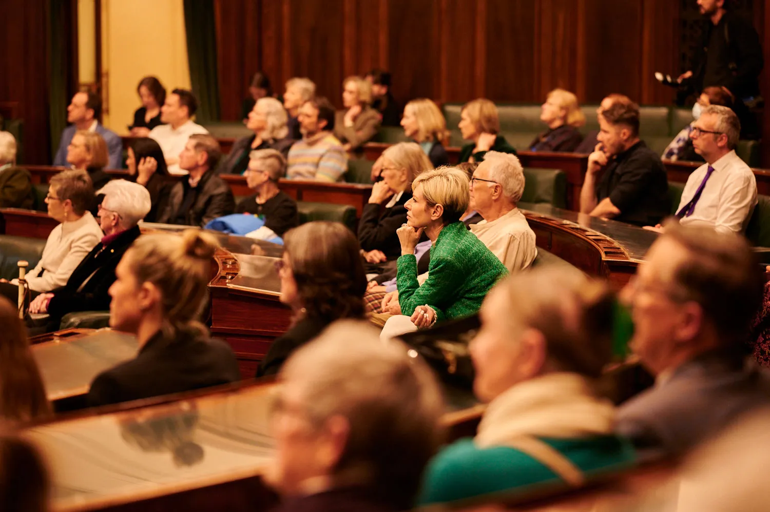 An audience sits on green leather benches in the House of Representatives at Old Parliament House. The room has wood panelled walls.