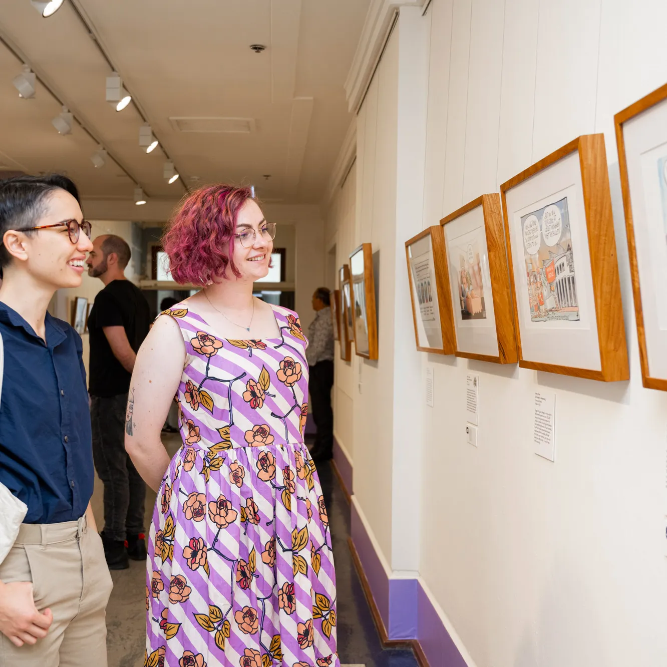 Two smiling people look at framed cartoons on the wall of an exhibition. 