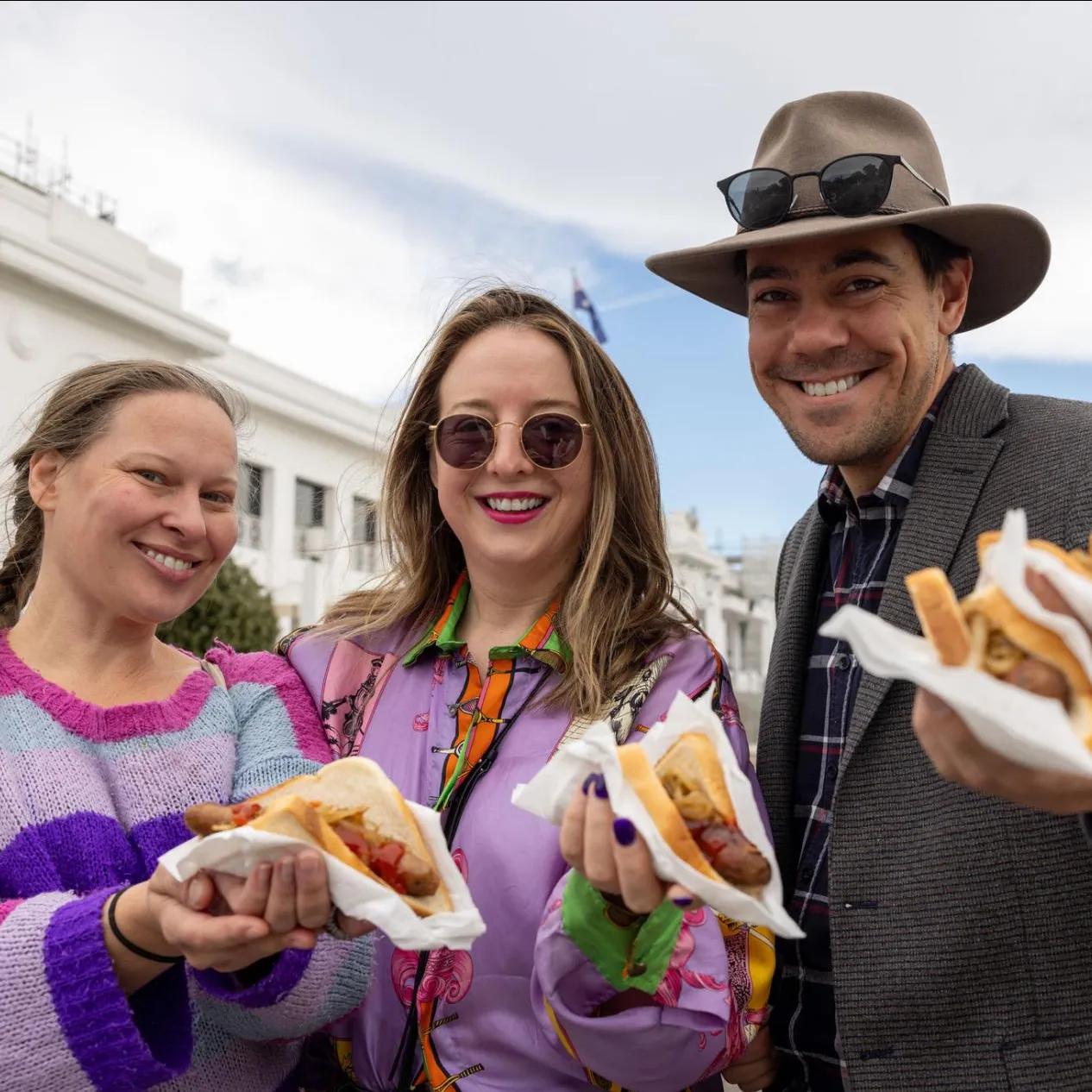 Two women wearing brightly coloured purple tops and a man in a jacket and hat stand in front of Old Parliament House holding sausage sandwiches.