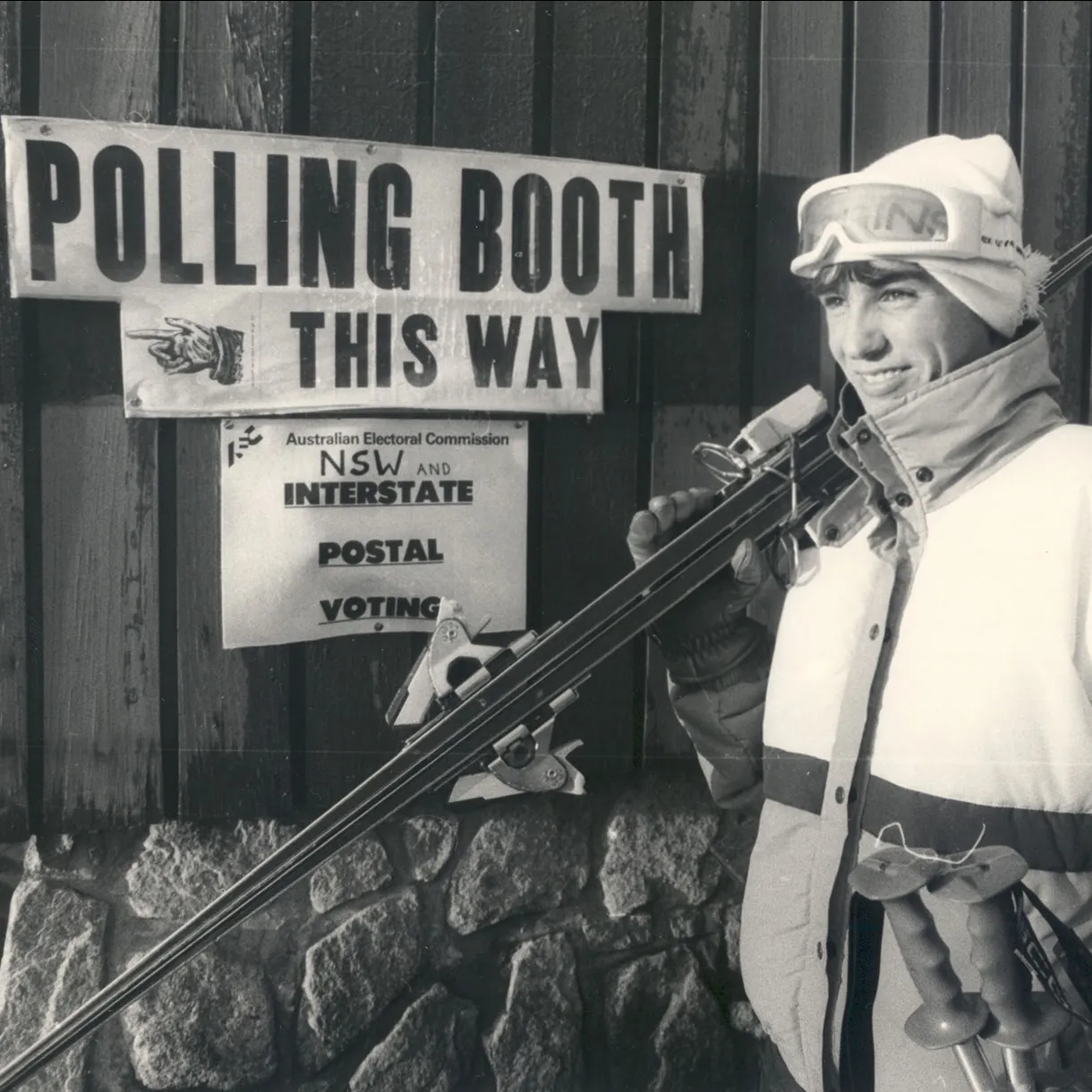 A man wearing vintage ski clothes and carrying skis stands outside a building with a 'Polling booth this way' sign.