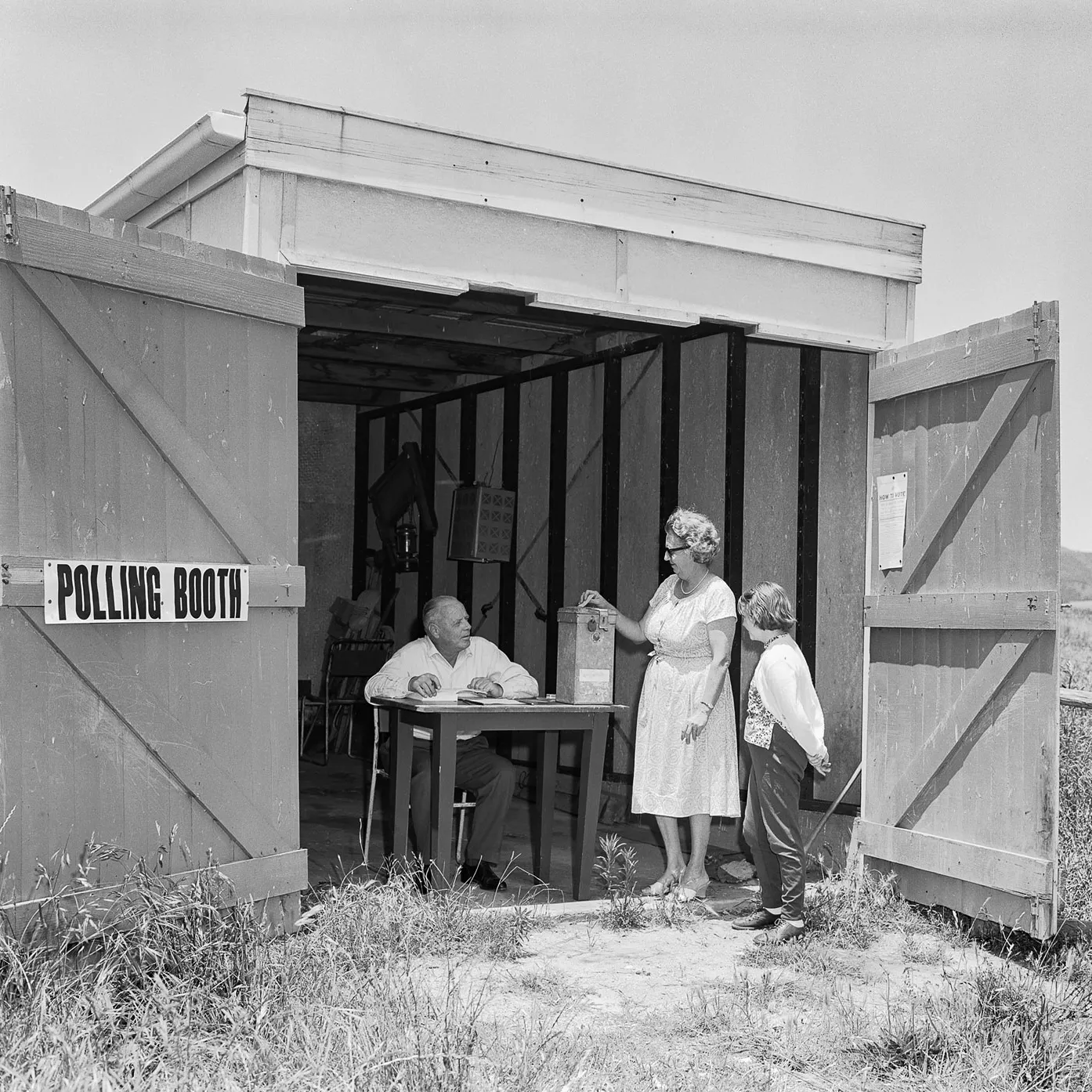 A woman puts a voting card in a ballot box at a shed in a paddock being used as a polling booth. A man sits at a table and a child looks on. 