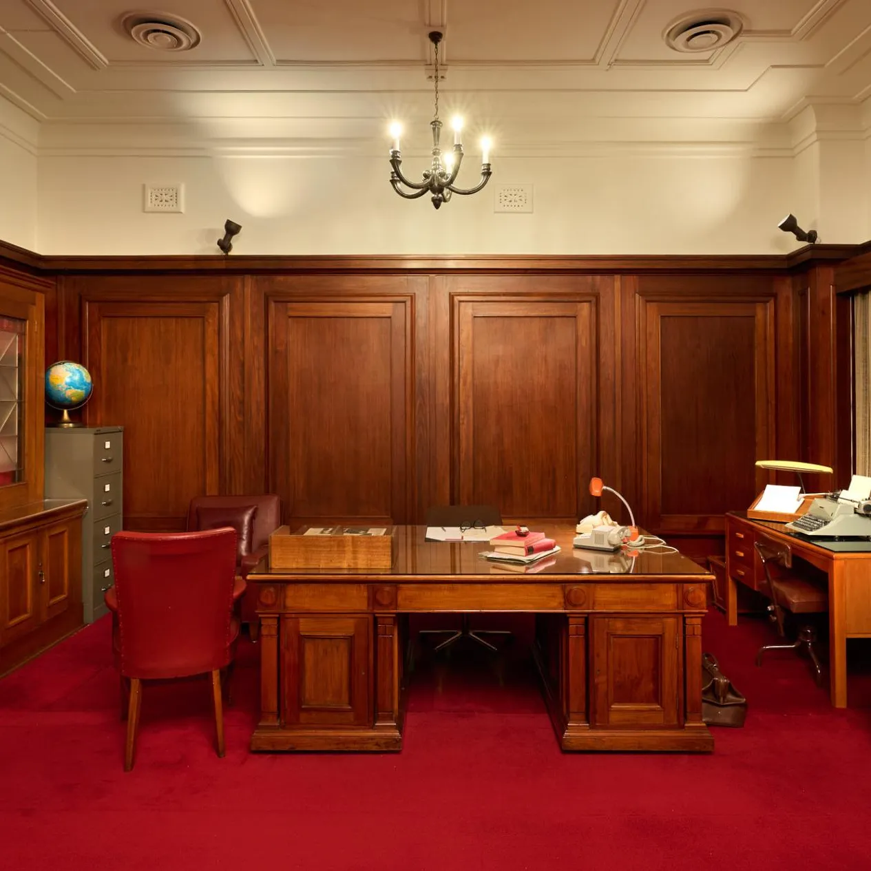 A wood-panelled room in Old Parliament House with two wooden desks, a wooden bookcase, red leather chairs and a red leather couch. The carpet is red.