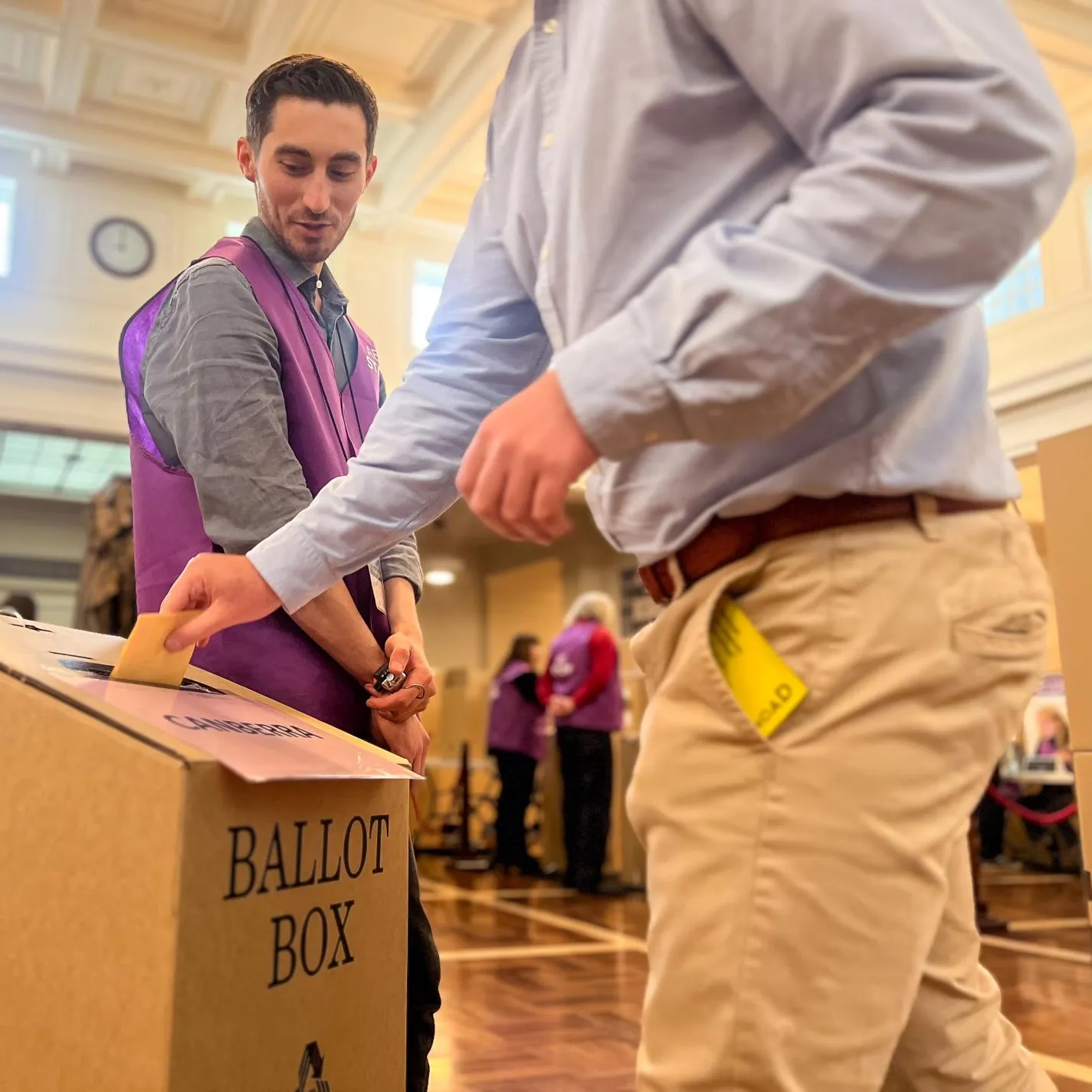 A voter puts a ballot paper in a cardboard ballot box while an AEC official dressed in a purple AEC vest looks on. They are in King's Hall in Old Parliament House.