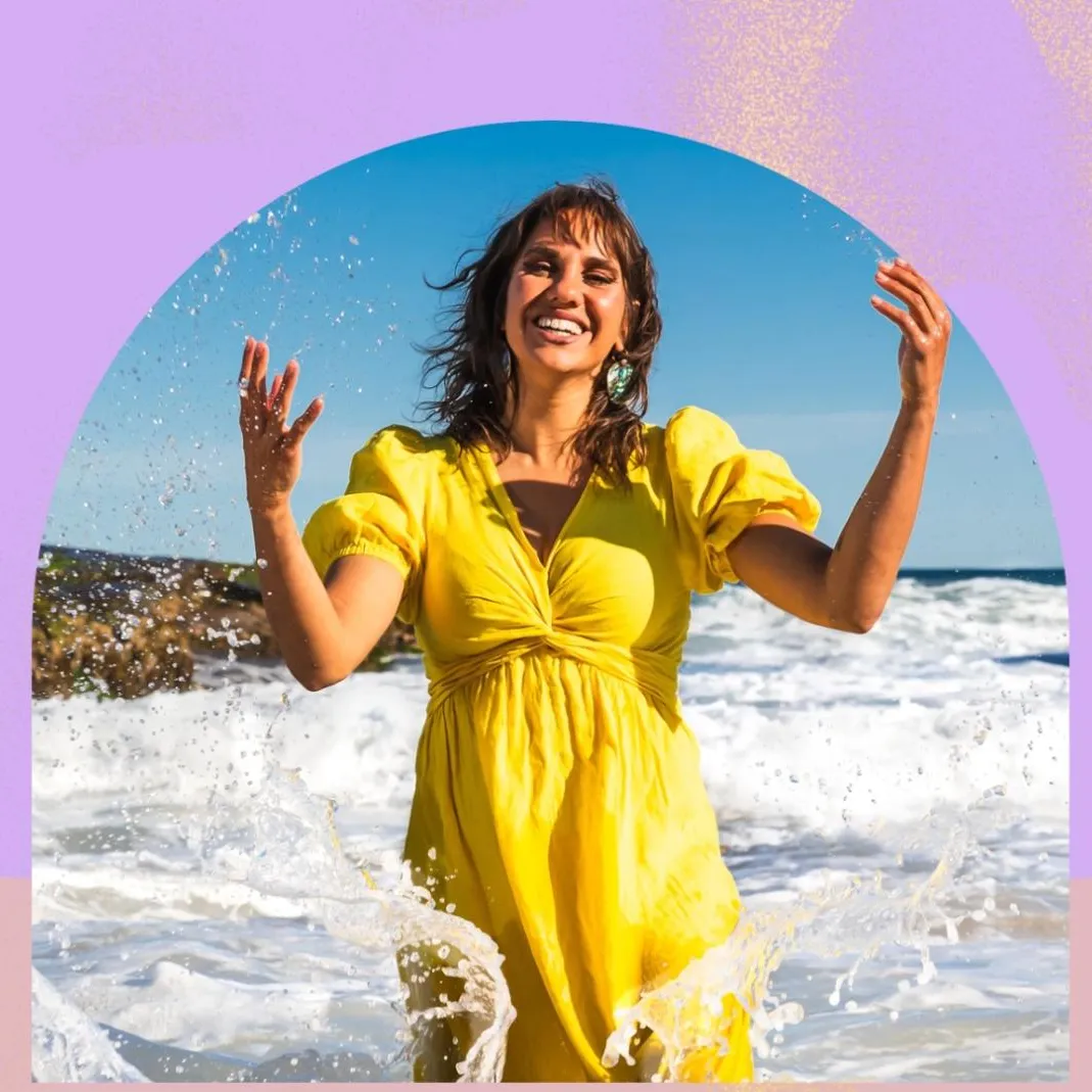 Kirli Saunders wears a yellow dress and stands in the waves on a beach.