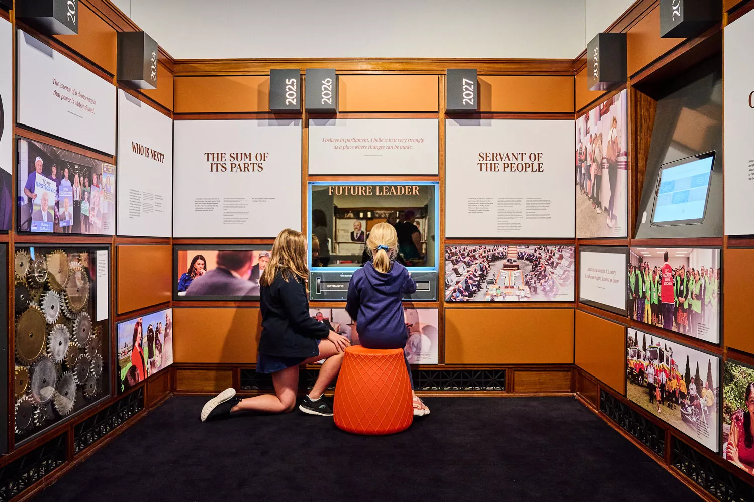 Two girls look into a mirror that says 'Future Leader' in an alcove suurrounded by exhibition panels with titles such as Servant of the people and dates like 2025, 2027. 