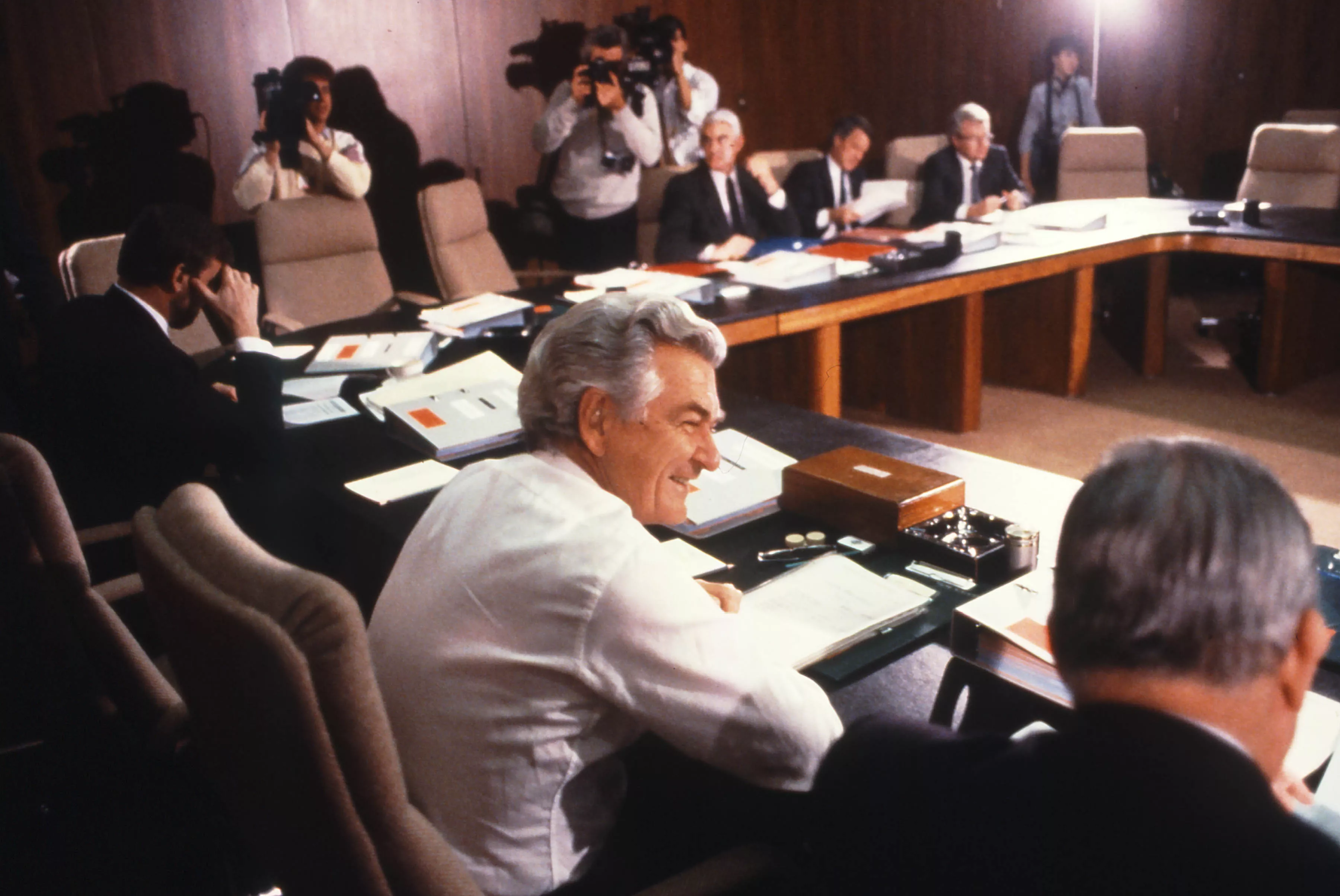 This colour photograph is taken from behind Deputy Prime Minister Lionel Bowen who is seated beside Prime Minister Bob Hawke. Both are dressed in standard business attire.  Other members of Cabinet can be seen sitting around the table although only half of the seats are filled. In the background are a photographer and two cameramen photographing and filming the cabinet meeting. 