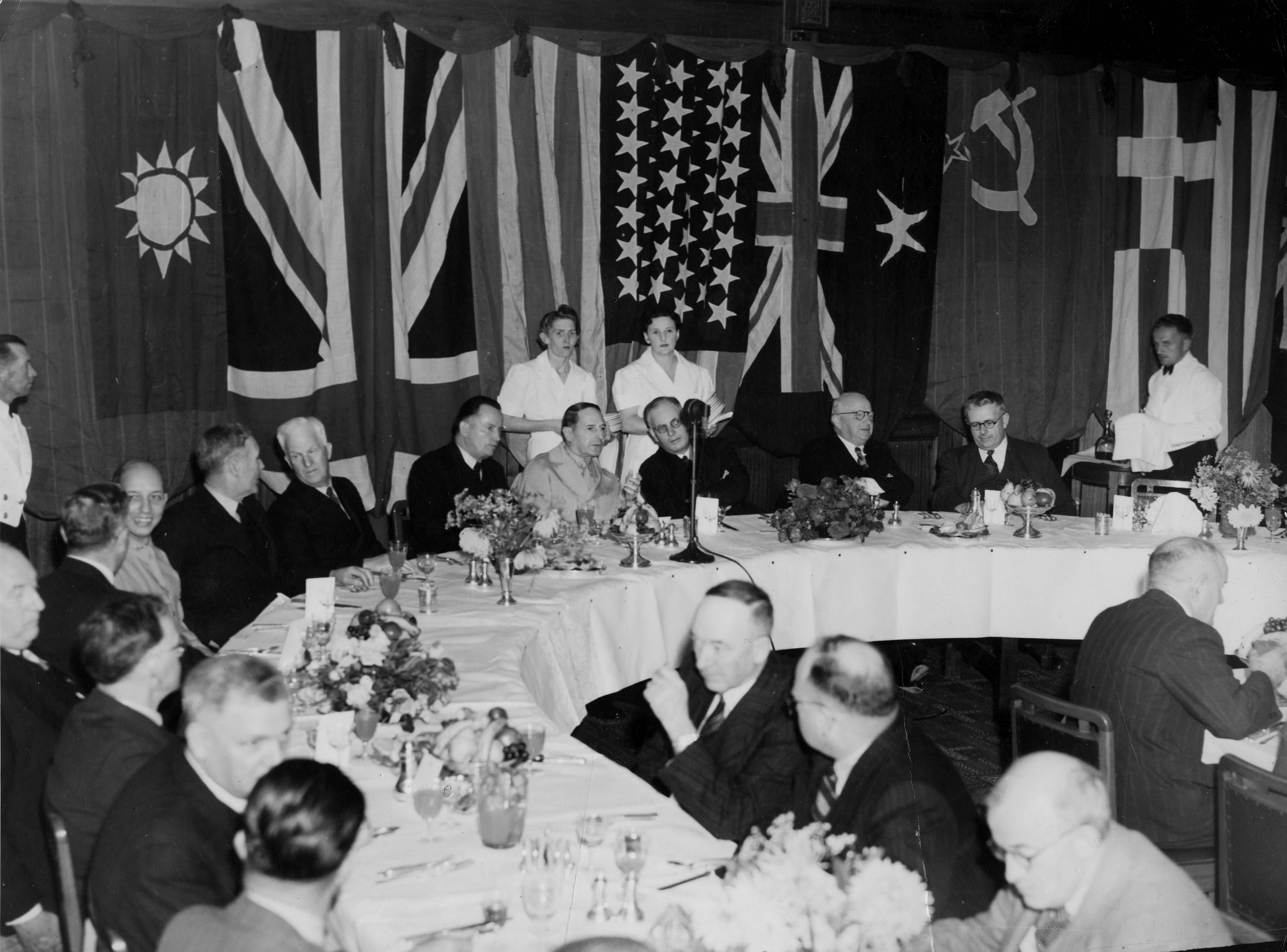 This black and white photograph shows a section of a dining table in the Members’ Dining Room with national flags hanging in the background. General Douglas MacArthur is seated at the head of the table with a microphone in front of him; he is between Deputy Leader of the ALP Frank Forde and Prime Minister John Curtin.  Other politicians are seated at the table which is set with white linen, silverware, glassware, crockery and flower arrangements. Uniformed waiters and waitresses stand behind the diners. 