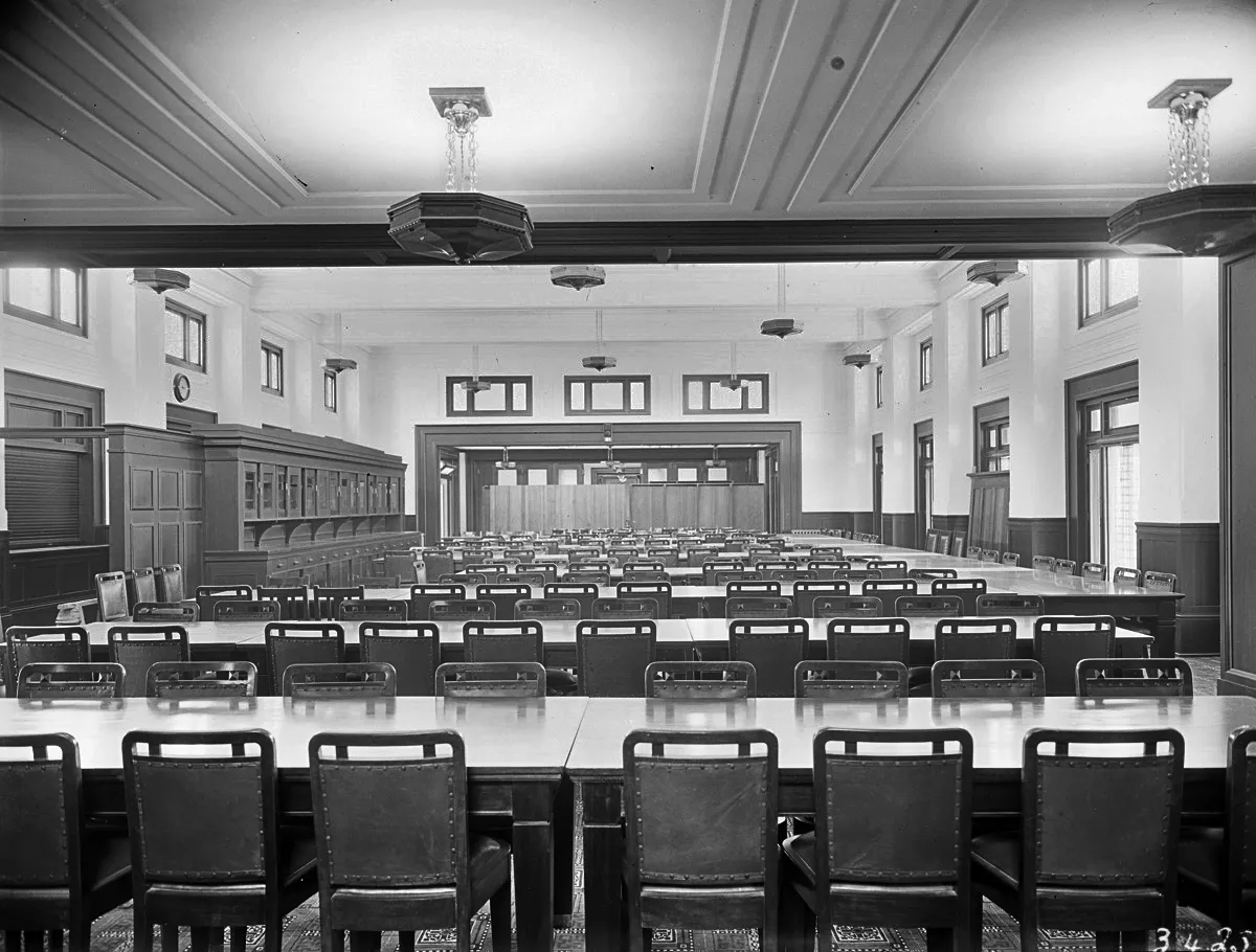 This black and white photograph looks along the length of the high-ceilinged Members’ Dining Room at Parliament House. Northern light floods in from windows and French doors on the right-hand wall. Rows of rectangular dining tables surrounded by hundreds of dining chairs span the width of the crowded room and on the left-hand wall is a timber servery. 