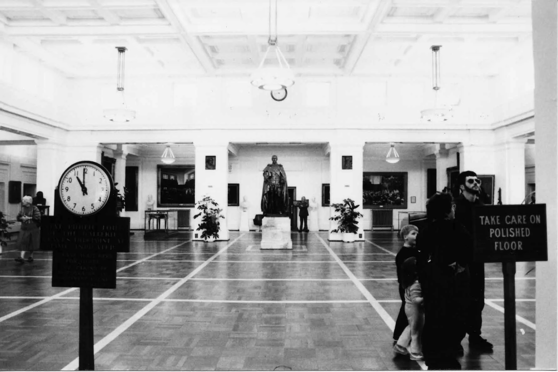 This black and white photograph of King’s Hall looks along the length of the room to the slightly larger than life bronze statue of King George the Fifth on a marble pedestal. King’s Hall is a large white room with a vaulted ceiling, celerestory windows, colonnades and a polished parquet floor.  The walls and columns feature artworks and portraits. You can see people wandering through the space including a family with young children and an older lady. In the foreground are two signs.