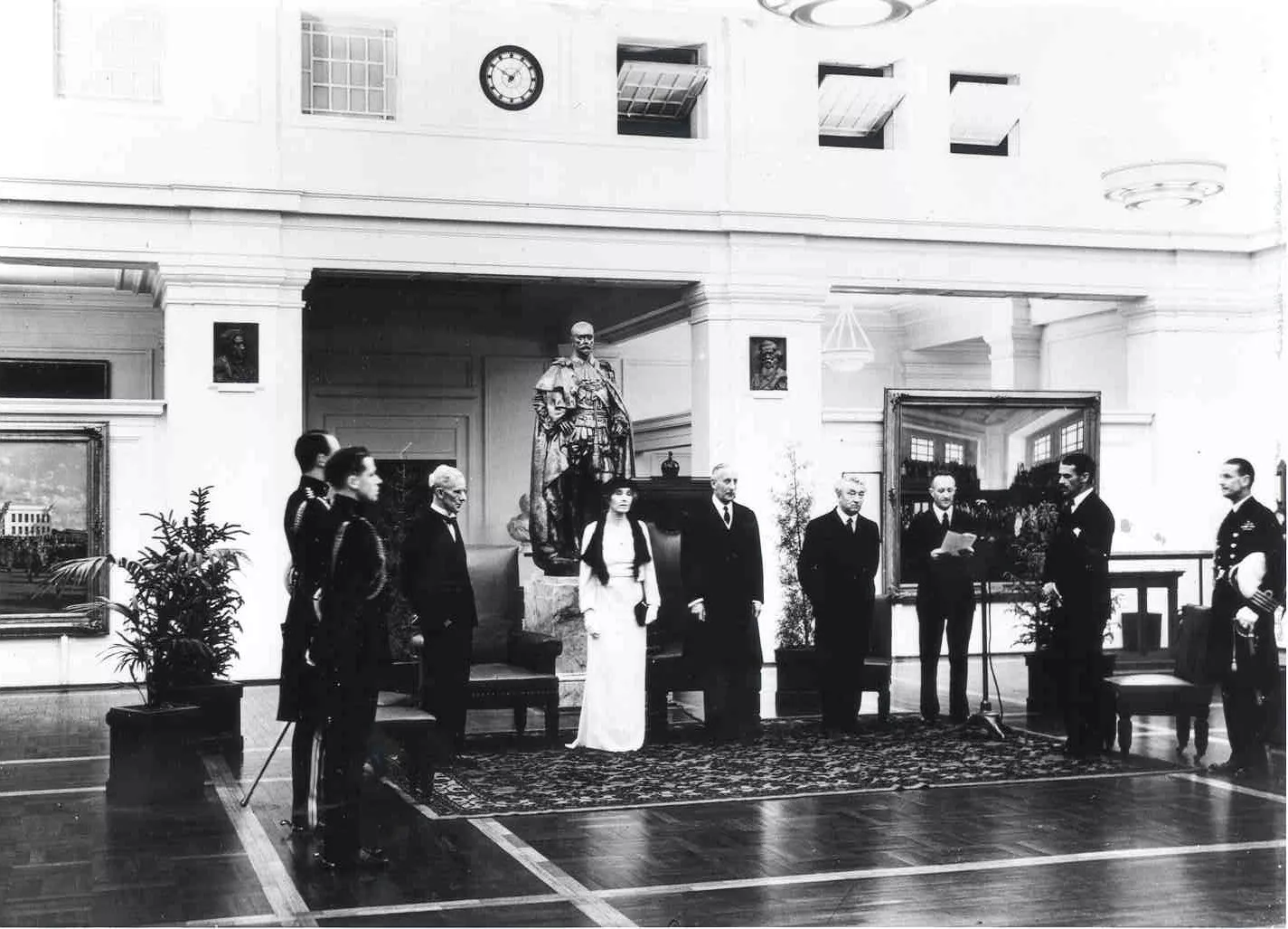 This black and white photograph shows a group of people standing at attention in King’s Hall while the announcement is read. The statue of George the Fifth looms over the group. Ther are seats behind the attendees,  including the Vice Regal Chair which would have been moved from the Senate Chamber. The men wear dark suits or military uniforms while the Governor-General’s wife, Lady Gowrie, wears a white formal dress, hat and gloves.