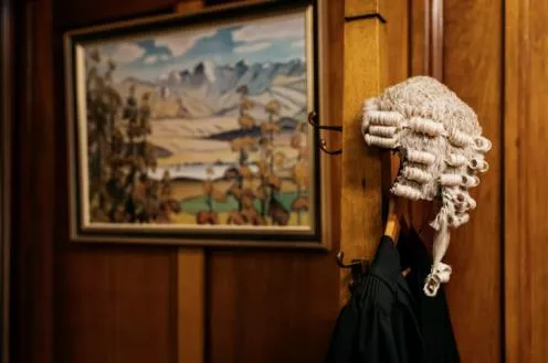 A wooden panelled wall with a framed piece of artwork. To the right of it stands a coat rack with a a jacket hanging from it and a judge's wig resting on top.