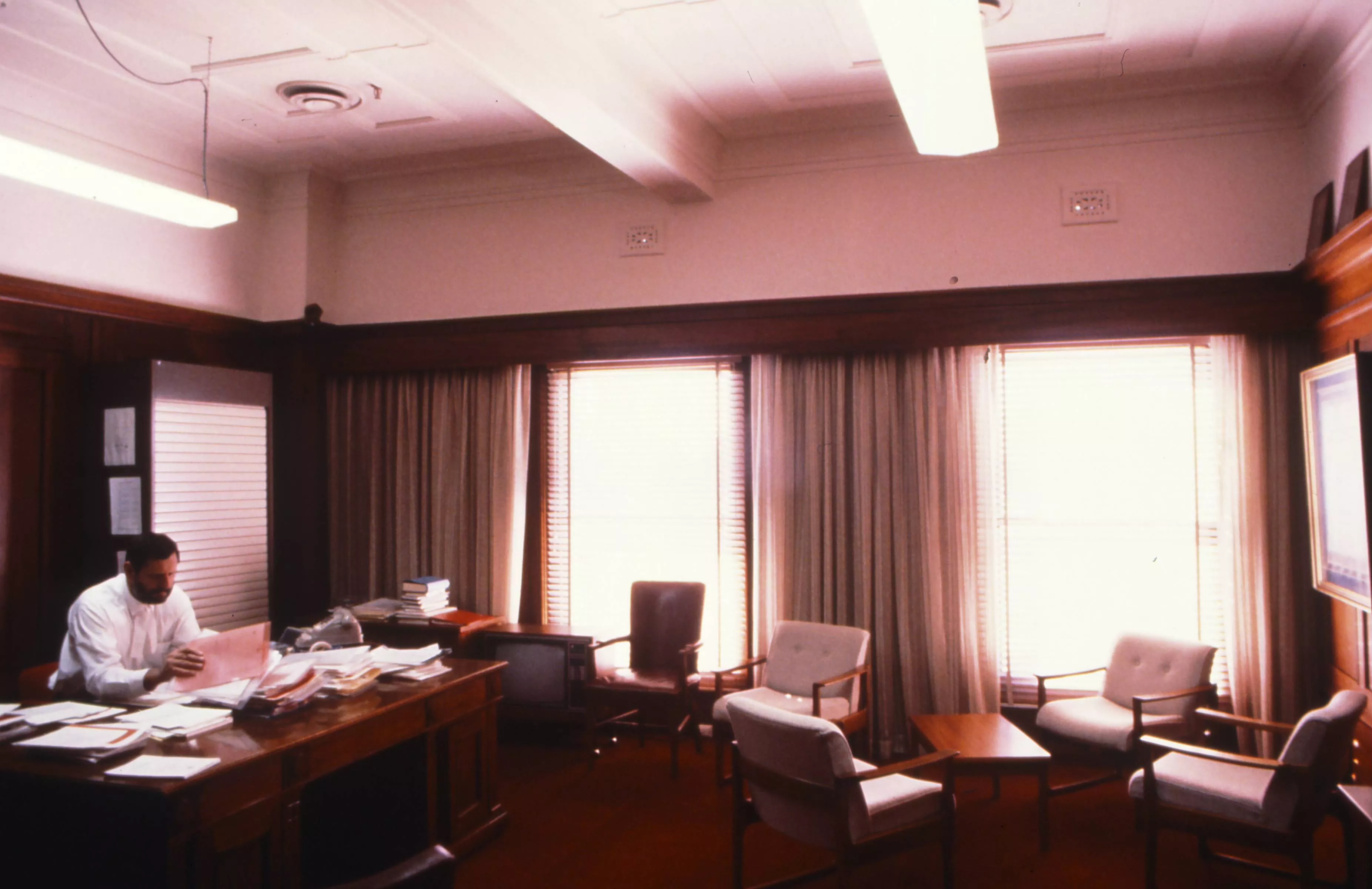 This colour photograph captures Harry Jenkins, the Clerk of the Senate, sitting at his desk in his office. The timber-panelled room is flooded with northern light from two large windows. The desktop is piled with papers and files and there is a grey telephone at Jenkins’ left hand. There is a tall filing cupboard and a sideboard piled high with papers and books in one corner. A bulky floor-mounted television sits alongside the sideboard. Four visitor chairs haphazardly surround a small coffee table and ther